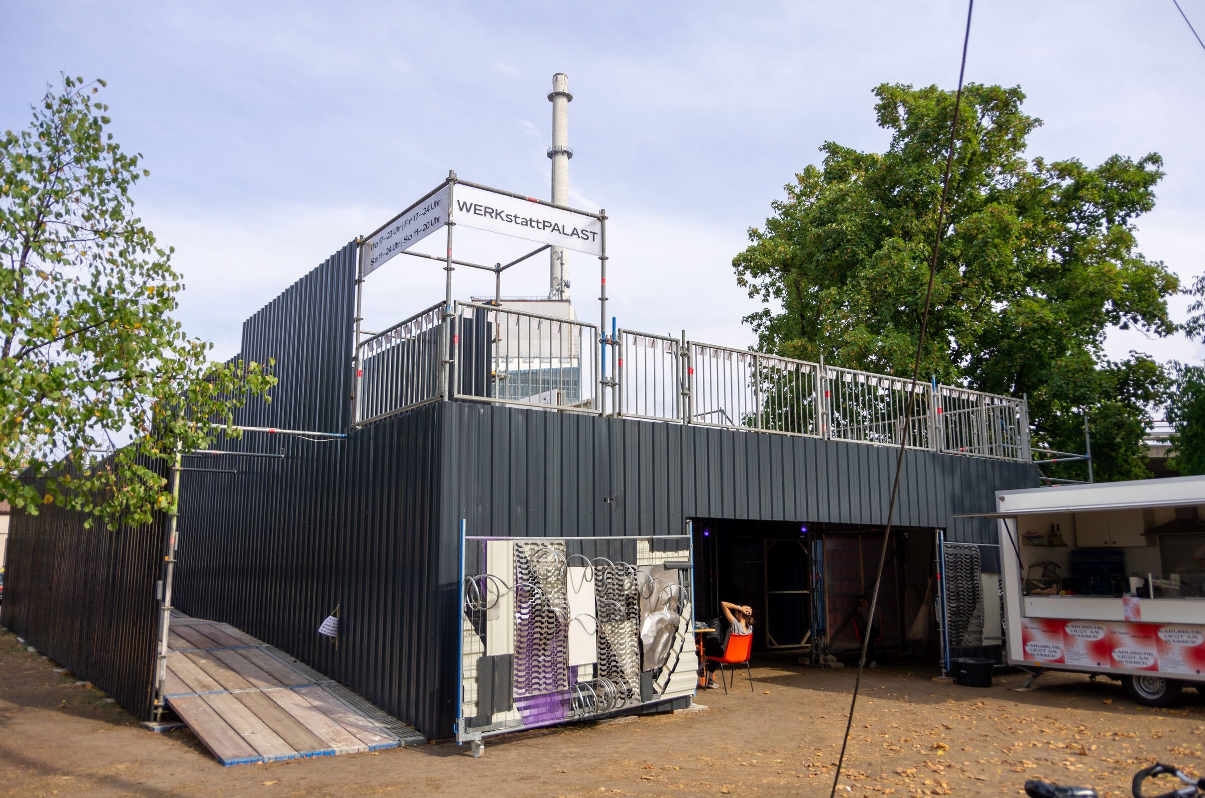 Außenansicht des dunklen Gerüstbaus mit Dachterrasse, Rampe und Bäumen am Rand.