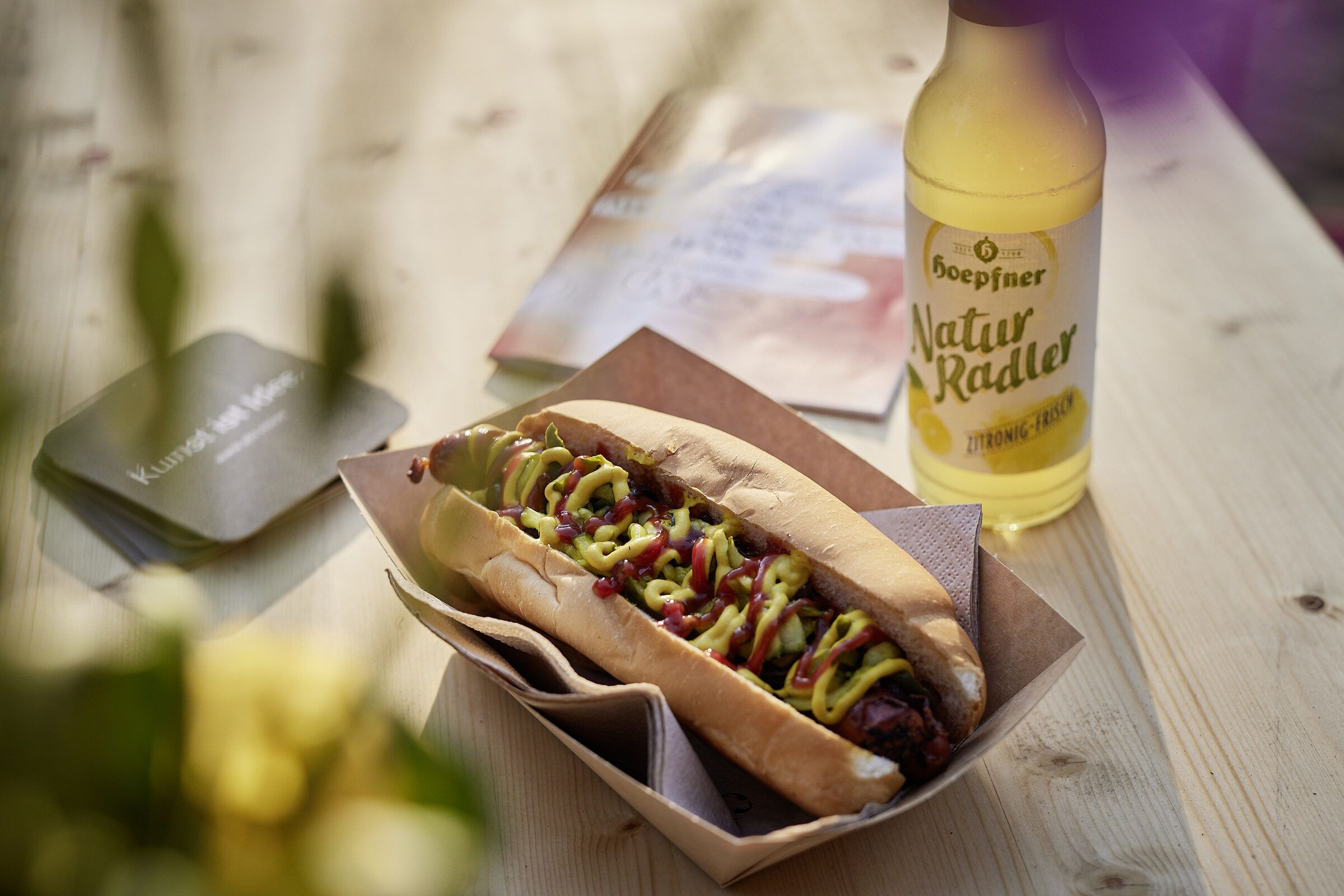 Ein Hotdog in einer Pappschale neben einer Flasche Naturradler auf einem Holztisch.
