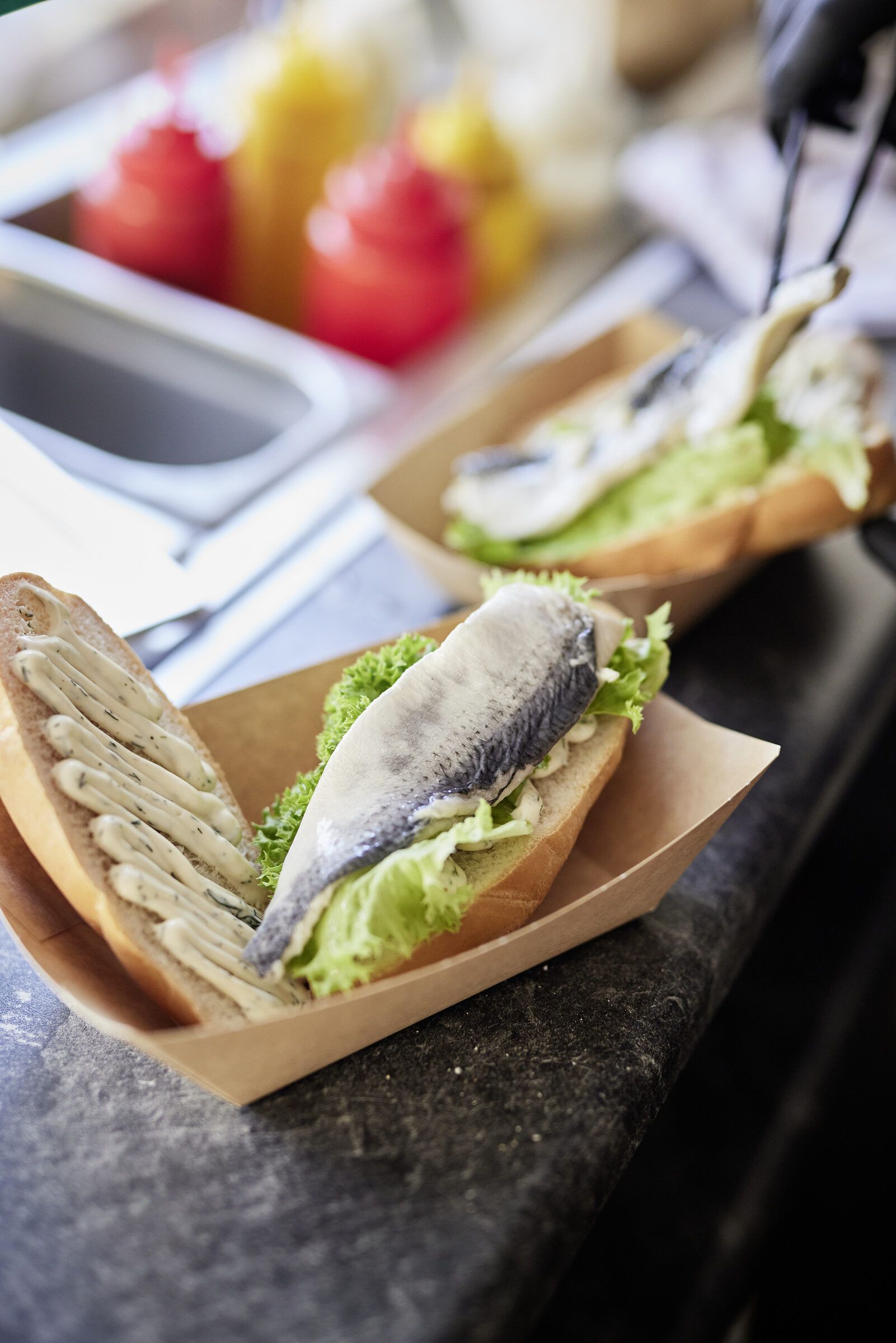 Ein belegtes Fischbrötchen in einer Pappschale auf einer Theke, daneben Obst.
