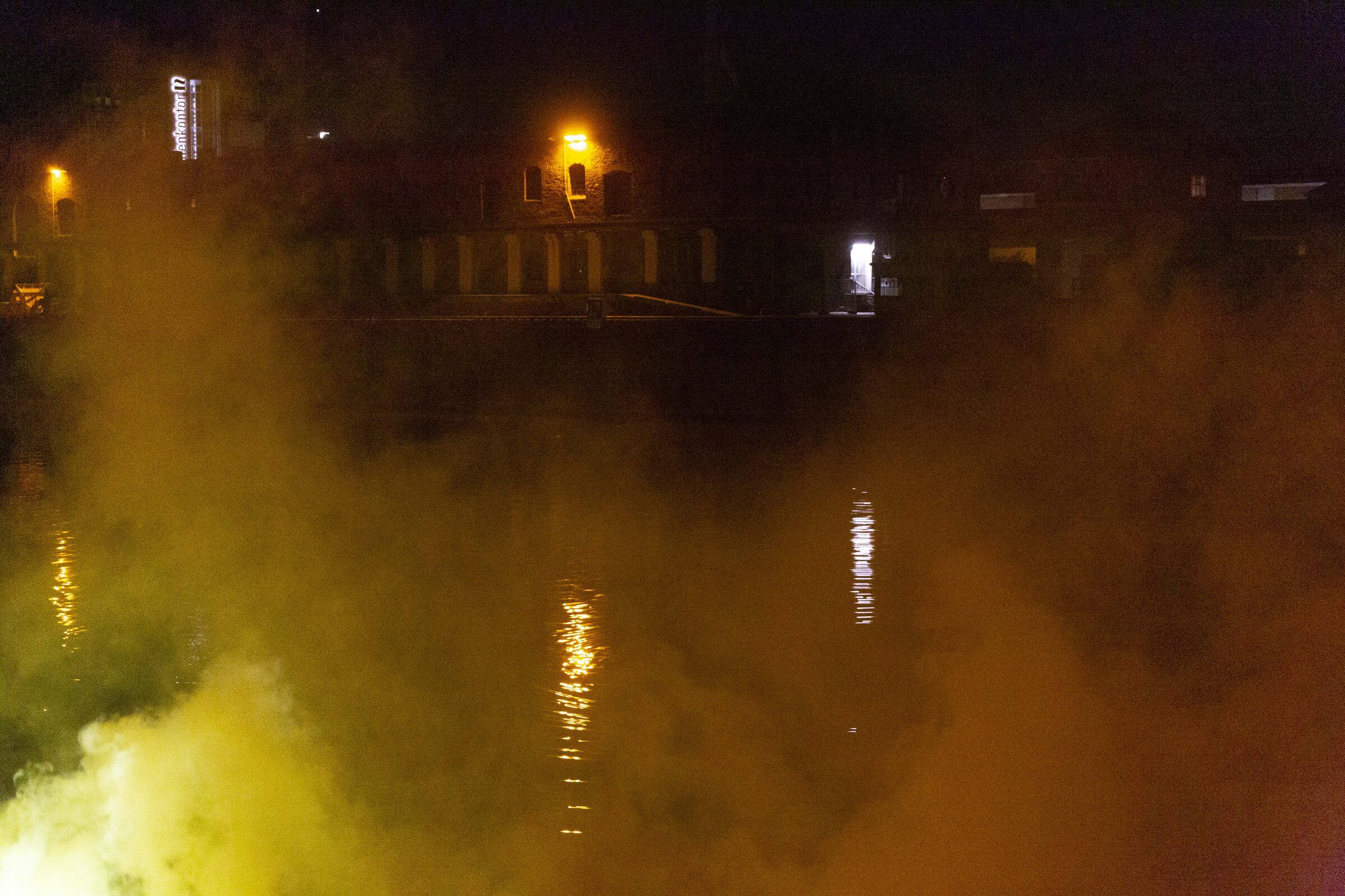 Gelber Rauch zieht nachts über das dunkle Hafenbecken, am Ufer brennen Lichter.