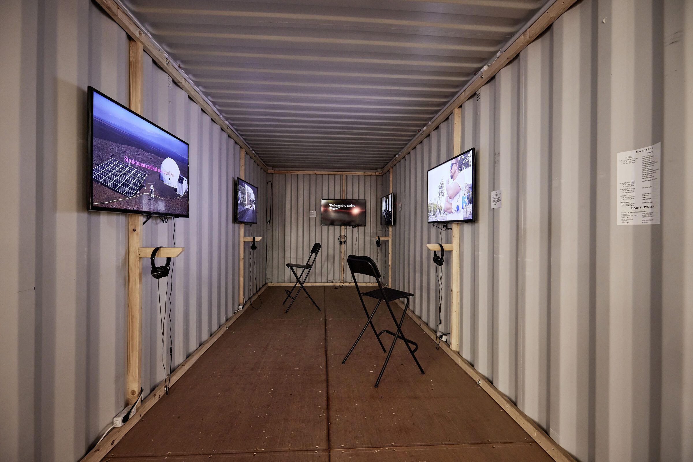 Container mit Bildschirmen an beiden Wänden und zwei Klappstühlen.