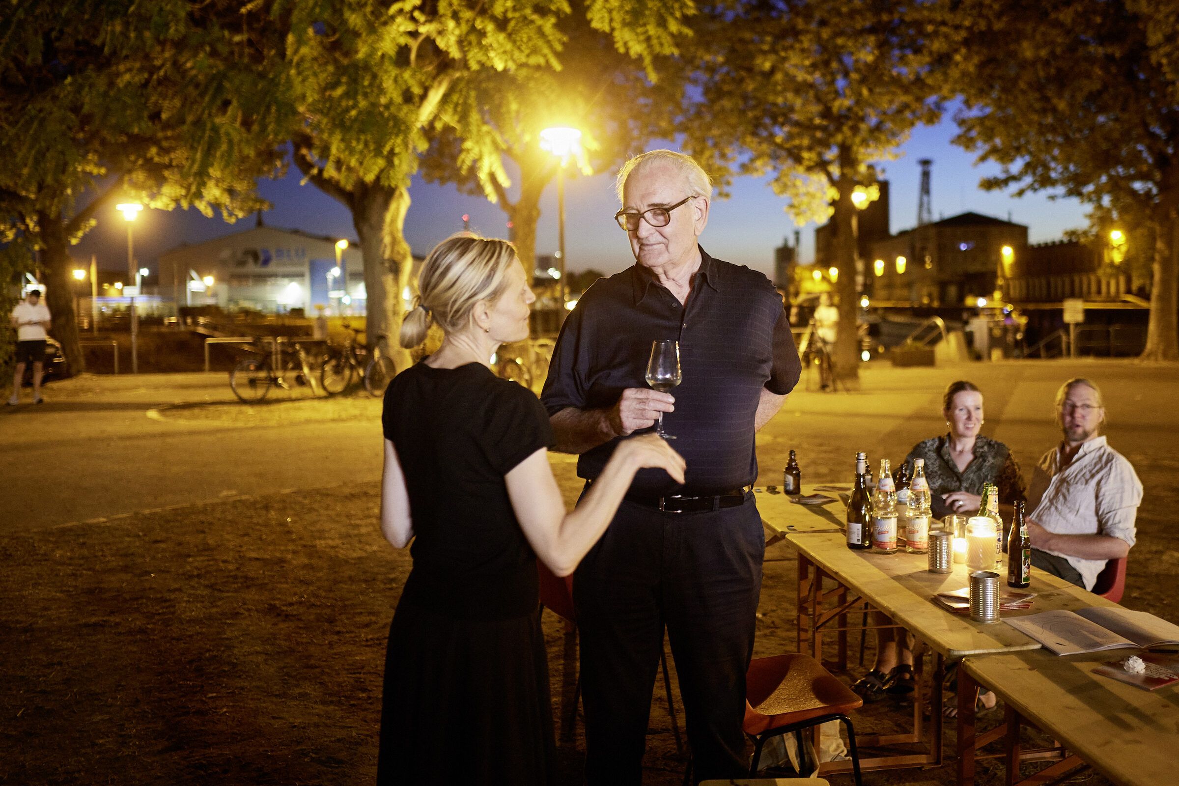 Zwei Gäste unterhalten sich nachts mit Weingläsern an der langen Tafel unter Laternenlicht.