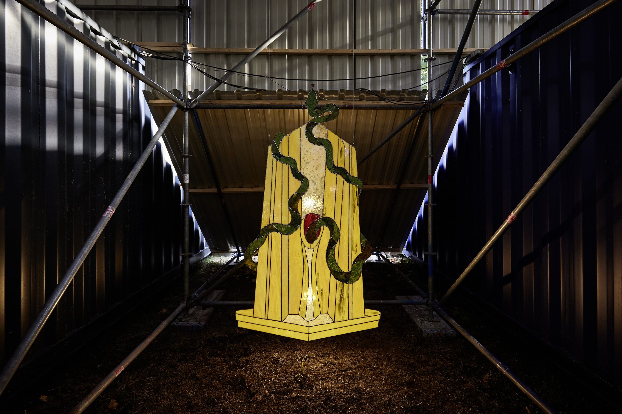 Ein leuchtendes gelbes Buntglasbild mit grüner Schlange hängt im dunklen Stahlraum.