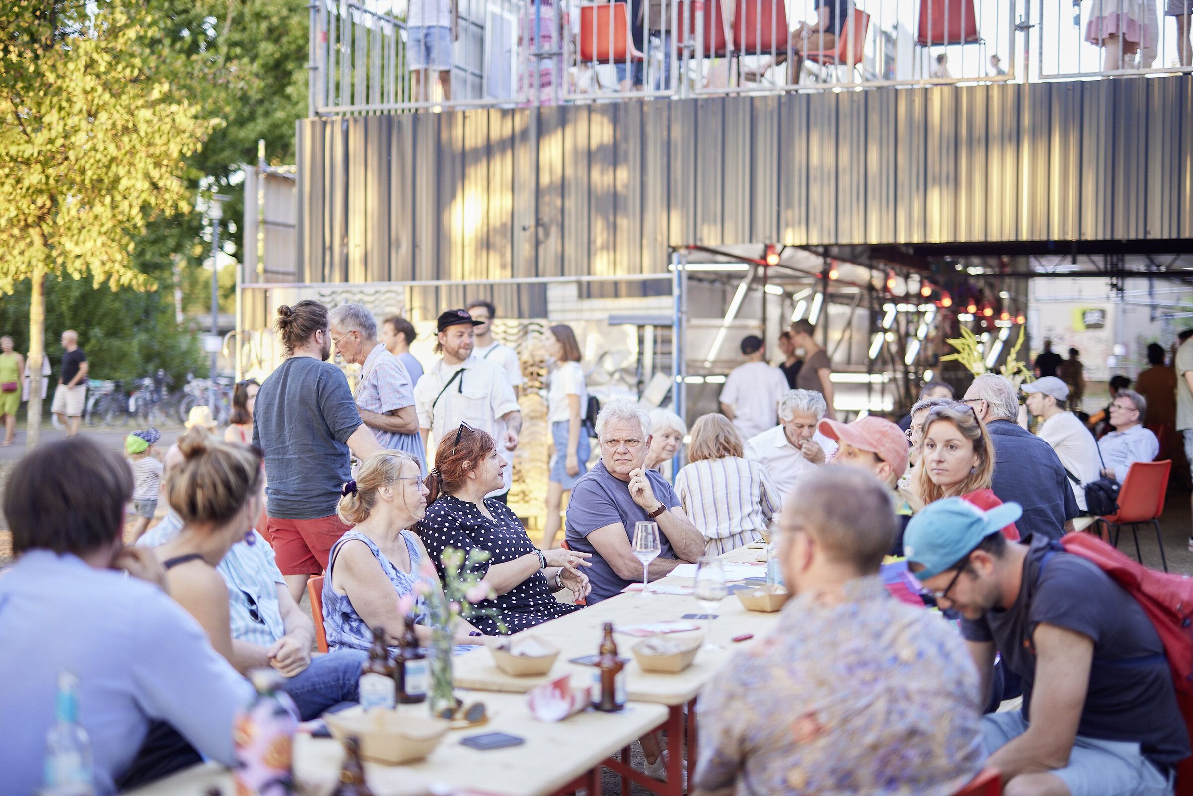 Voll besetzte lange Tafel vor dem Gerüstbau, Menschen essen, trinken und unterhalten sich.