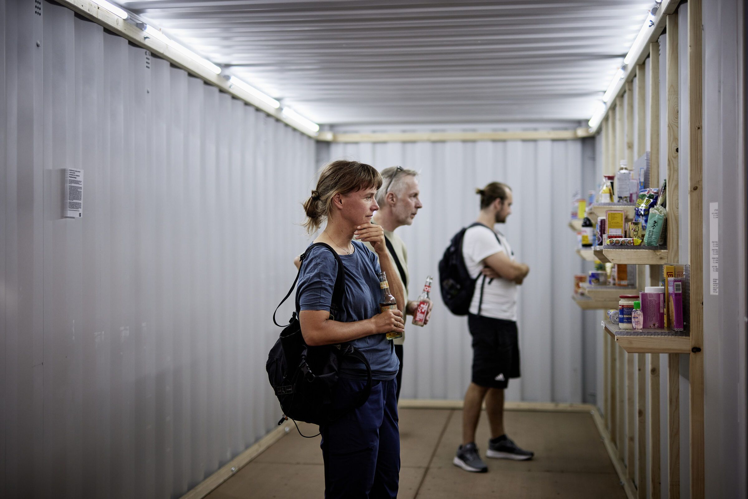 Drei Besucher:innen betrachten im schmalen Container die Regale mit Produkten.